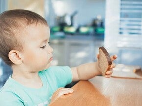Niño pequeño sentado en una mesa de madera en la cocina, con camiseta verde, sosteniendo una rebanada de pan integral.
