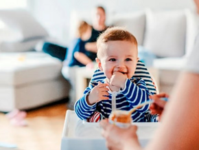 Bebé listo para comer alimentos sólidos, sonriendo y sentado en una silla alta mientras la mamá le pasa una cucharadita con compota.