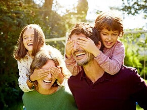 Familia de cuatro disfrutando de un día soleado al aire libre. Dos adultos llevan a dos niños seguros y confiados sobre sus hombros, rodeados de árboles y vegetación