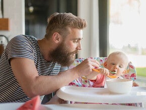 Hombre con barba alimentando papilla a un bebé en una silla alta. En la mesa hay un bol blanco.