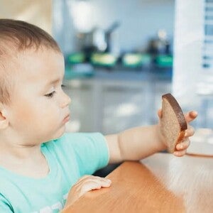 Niño pequeño sentado en una mesa de madera en la cocina, con camiseta verde, sosteniendo una rebanada de pan integral.