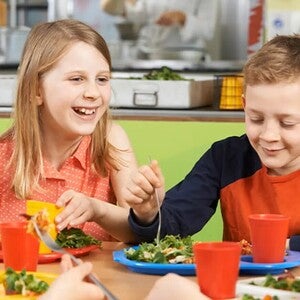 Niños comiendo en una cafetería escolar con platos de ensalada y pasta.