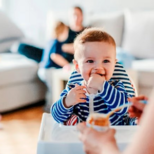 ¿Tu bebé está listo para comenzar con sólidos? Bebé listo para comer alimentos sólidos, sonriendo y sentado en una silla alta mientras la mamá le pasa una cucharadita con compota.
