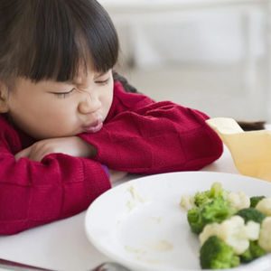 ¿Es normal que mi bebé no quiera comer? Niña pequeña con cara de disgusto, en frente tiene un plato con brócoli y coliflor.