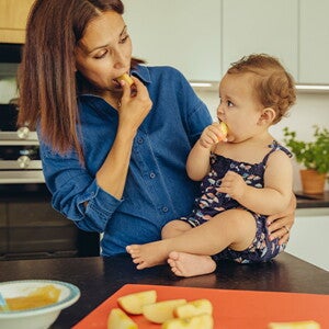 Identifica cuáles son las frutas ideales para iniciar esta nueva etapa de tu bebé. Mamá y bebé comiendo fruta en la cocina.