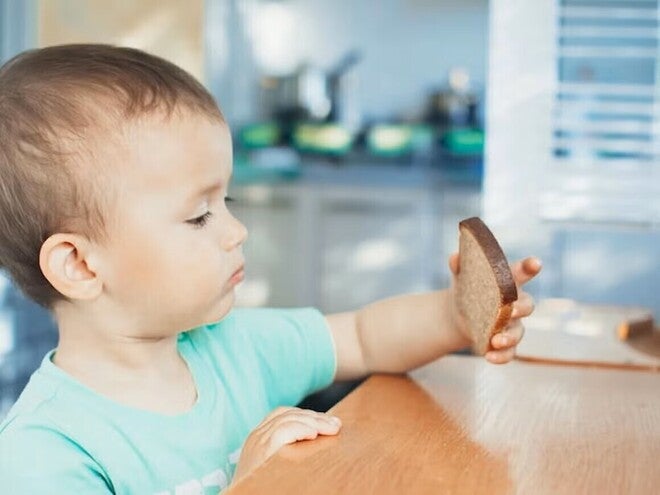 Niño pequeño sentado en una mesa de madera en la cocina, con camiseta verde, sosteniendo una rebanada de pan integral.