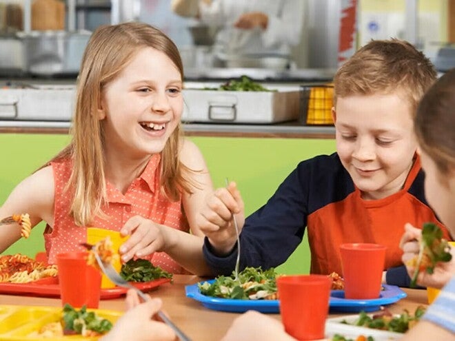 Niños comiendo en una cafetería escolar con platos de ensalada y pasta.