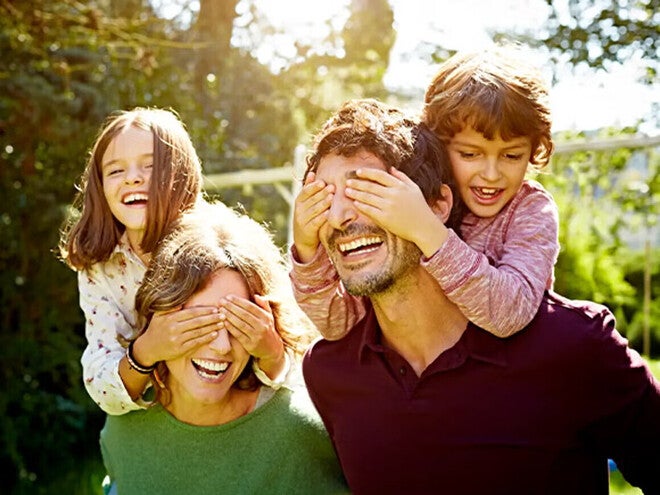 Familia de cuatro disfrutando de un día soleado al aire libre. Dos adultos llevan a dos niños seguros y confiados sobre sus hombros, rodeados de árboles y vegetación