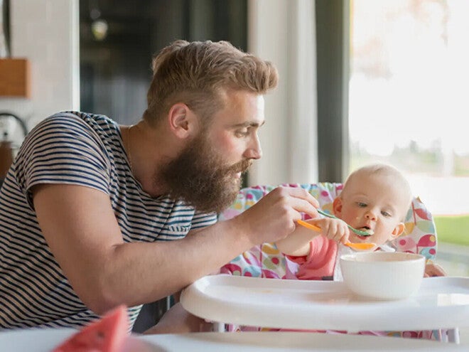 Hombre con barba alimentando papilla a un bebé en una silla alta. En la mesa hay un bol blanco.